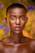 A model with a shaved head and glowing skin is centered in the image, wearing striking makeup with vibrant colors around the eyes, including red and yellow eyelids with precise eyeliner. The background features soft, colorful bokeh lights in shades of blue, orange, and green, creating an artistic and dreamy effect.