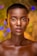 A model with a shaved head and glowing skin is centered in the image, wearing striking makeup with vibrant colors around the eyes, including red and yellow eyelids with precise eyeliner. The background features soft, colorful bokeh lights in shades of blue, orange, and green, creating an artistic and dreamy effect.