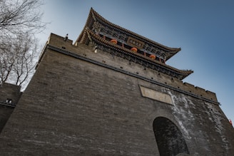 A traditional Chinese architectural structure with ornate roofing and intricate details stands against a blue sky. The large, fortified gate is made of brick and features decorative elements, with a sign in Chinese characters. Leafless trees can be seen to the side of the structure.