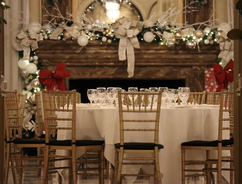 A round wedding table laid with white linen, glasses and gold chairs in front of a fireplace dressed with garland, ribbon and fairy lights