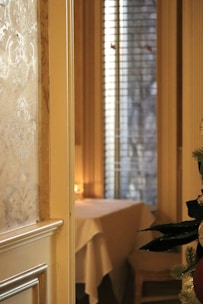 An elegant interior setting with a softly lit, patterned wall beside a window with vertical blinds. In the foreground, part of a decorated Christmas tree with ornaments is visible. A table covered with a white tablecloth sits in the background, illuminated by warm, ambient lighting.