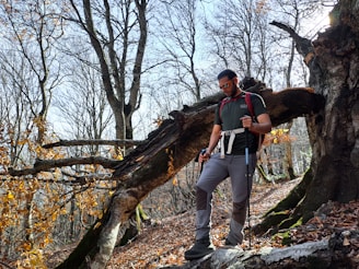 A man wearing hiking gear, including sunglasses and a backpack, is using trekking poles while navigating a forest path. The surrounding area is filled with tall, leafless trees and a large fallen tree trunk, with some remaining autumn leaves adding a touch of color.