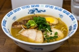 A bowl of ramen is filled with rich broth, topped with slices of tender pork, half a soft-boiled egg, bamboo shoots, and fresh green onions. The bowl has blue decorative patterns on its rim.