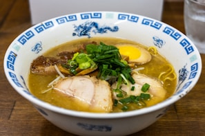 A bowl of ramen is filled with rich broth, topped with slices of tender pork, half a soft-boiled egg, bamboo shoots, and fresh green onions. The bowl has blue decorative patterns on its rim.