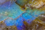A close-up of a mineral or rock surface with an iridescent, multi-colored sheen, displaying shades of blue, green, and purple. The surface appears rough and textured with various small lines and specks scattered throughout.