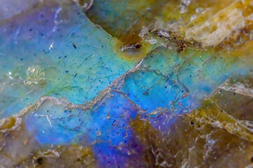 A close-up of a mineral or rock surface with an iridescent, multi-colored sheen, displaying shades of blue, green, and purple. The surface appears rough and textured with various small lines and specks scattered throughout.