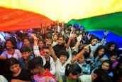 A large group of people, mostly young, gather closely together beneath a large rainbow-colored fabric. Many individuals are smiling, raising their hands, and displaying an overall sense of joy and unity. The vibrant fabric overhead features visible stripes of yellow, orange, red, green, and blue.