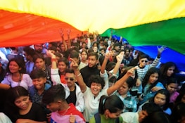A large group of people, mostly young, gather closely together beneath a large rainbow-colored fabric. Many individuals are smiling, raising their hands, and displaying an overall sense of joy and unity. The vibrant fabric overhead features visible stripes of yellow, orange, red, green, and blue.
