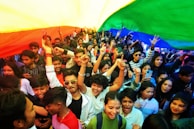 A large crowd of people celebrating under a rainbow flag, with many individuals raising their hands and showing enthusiasm. The scene is vibrant and full of young people expressing joy in a unified gathering.