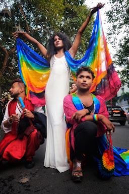 A person in a white dress stands with arms outstretched holding a colorful rainbow fabric behind them. Two other individuals, adorned in vibrant clothing with rainbow motifs, are kneeling on either side, exuding a sense of pride and celebration. The background features trees and a street scene.