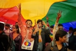 A group of people gathered under a large rainbow flag, smiling and celebrating. One person is holding a sign that says 'Born This Way'. Many of the participants have colorful clothing and accessories, and some have painted rainbow flags on their faces.