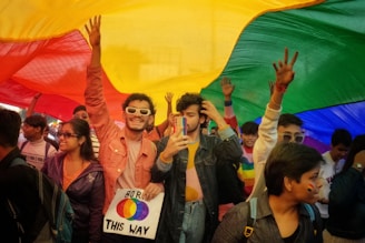 A group of people gathered under a large rainbow flag, smiling and celebrating. One person is holding a sign that says 'Born This Way'. Many of the participants have colorful clothing and accessories, and some have painted rainbow flags on their faces.