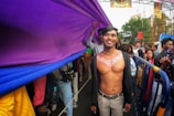 A shirtless person with decorative body paint stands smiling at a lively outdoor event. They are under a large, colorful purple and blue fabric banner being carried by others. There is a diverse crowd behind, some of whom hold the banner. Metal barriers line the street, and pride signs are visible in the background.