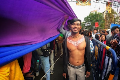 A shirtless person with decorative body paint stands smiling at a lively outdoor event. They are under a large, colorful purple and blue fabric banner being carried by others. There is a diverse crowd behind, some of whom hold the banner. Metal barriers line the street, and pride signs are visible in the background.
