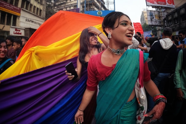 A lively crowd is gathered on a street for a parade or march. At the forefront, a person in colorful attire, including a turquoise sari and traditional jewelry, smiles joyfully. Behind them, a large rainbow flag is prominently displayed, held by a participant holding a smartphone. Surroundings include urban buildings, signage, and more people engaged in the celebration.