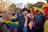 A group of people are gathered in a street, celebrating with rainbow flags and colorful clothing. There is a festive and energetic atmosphere. One person is holding a sign that references a 'gay agenda.' Buildings and trees can be seen in the background, with a city setting.