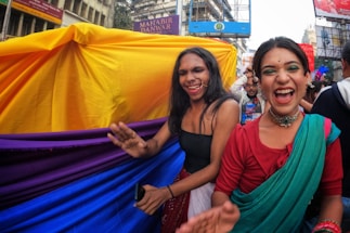 Two people wearing vibrant clothing and accessories are smiling and enjoying themselves in a lively outdoor setting. A colorful fabric canopy with bright yellow, purple, and blue shades sways behind them. The scene has an energetic and festive atmosphere, with other people visible in the background, participating in the celebration.