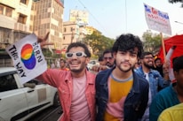 A crowd of people is seen participating in a colorful outdoor event. Two men in the foreground are smiling and holding signs supporting LGBTQ rights, one with the rainbow flag colors and 'Born This Way' written on it. The group is surrounded by buildings, trees, and clear skies, creating a lively and cheerful atmosphere.