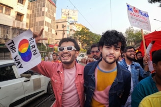 A crowd of people is seen participating in a colorful outdoor event. Two men in the foreground are smiling and holding signs supporting LGBTQ rights, one with the rainbow flag colors and 'Born This Way' written on it. The group is surrounded by buildings, trees, and clear skies, creating a lively and cheerful atmosphere.