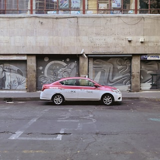 A red and white taxi is parked in front of a building with metal shutters that display graffiti art. The street is empty and the building appears to be old and slightly worn.
