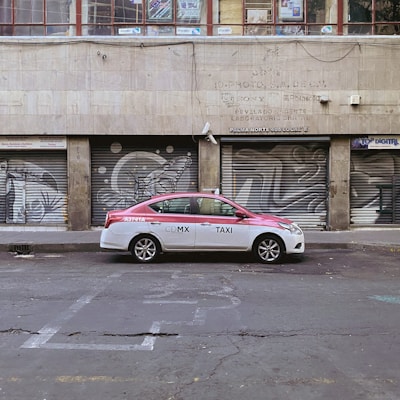 A red and white taxi is parked in front of a building with metal shutters that display graffiti art. The street is empty and the building appears to be old and slightly worn.