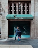 Two people stand in front of a closed jewelry store with a green awning labeled 'JOYERÍA'. The street is made of tiled concrete, and the building has a stone facade. There are metal bars on a window above the awning, and electrical wires run along the wall.