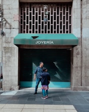Two people stand in front of a closed jewelry store with a green awning labeled 'JOYERÍA'. The street is made of tiled concrete, and the building has a stone facade. There are metal bars on a window above the awning, and electrical wires run along the wall.