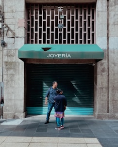 Two people stand in front of a closed jewelry store with a green awning labeled 'JOYERÍA'. The street is made of tiled concrete, and the building has a stone facade. There are metal bars on a window above the awning, and electrical wires run along the wall.