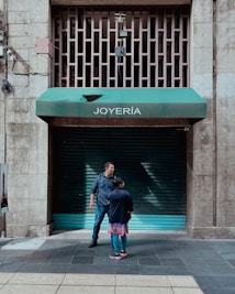 Two people stand in front of a closed jewelry store with a green awning labeled 'JOYERÍA'. The street is made of tiled concrete, and the building has a stone facade. There are metal bars on a window above the awning, and electrical wires run along the wall.