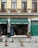 A storefront with a green awning that reads 'Artículos Religiosos y Artesanías' is displayed. The façade features tan brickwork with tall wooden-framed windows and black iron railings above. The entrance is flanked by stone columns, and a variety of religious and artisan items can be seen inside the shop. In front of the store, potted plants and cement blocks are arranged on a paved street.