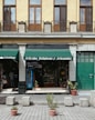 A storefront with a green awning that reads 'Art&iacute;culos Religiosos y Artesan&iacute;as' is displayed. The fa&ccedil;ade features tan brickwork with tall wooden-framed windows and black iron railings above. The entrance is flanked by stone columns, and a variety of religious and artisan items can be seen inside the shop. In front of the store, potted plants and cement blocks are arranged on a paved street.