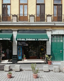 A storefront with a green awning that reads 'Art&iacute;culos Religiosos y Artesan&iacute;as' is displayed. The fa&ccedil;ade features tan brickwork with tall wooden-framed windows and black iron railings above. The entrance is flanked by stone columns, and a variety of religious and artisan items can be seen inside the shop. In front of the store, potted plants and cement blocks are arranged on a paved street.
