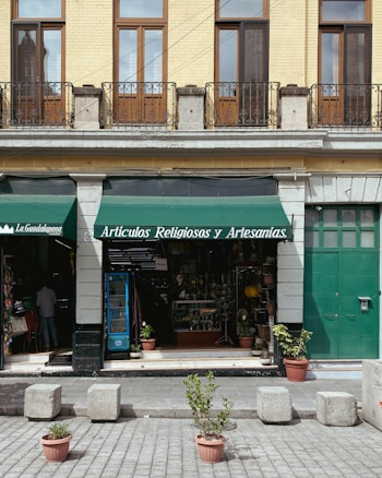 A storefront with a green awning that reads 'Art&iacute;culos Religiosos y Artesan&iacute;as' is displayed. The fa&ccedil;ade features tan brickwork with tall wooden-framed windows and black iron railings above. The entrance is flanked by stone columns, and a variety of religious and artisan items can be seen inside the shop. In front of the store, potted plants and cement blocks are arranged on a paved street.