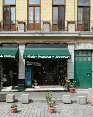 A storefront with a green awning that reads 'Art&iacute;culos Religiosos y Artesan&iacute;as' is displayed. The fa&ccedil;ade features tan brickwork with tall wooden-framed windows and black iron railings above. The entrance is flanked by stone columns, and a variety of religious and artisan items can be seen inside the shop. In front of the store, potted plants and cement blocks are arranged on a paved street.