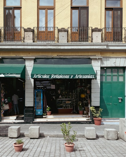 A storefront with a green awning that reads 'Art&iacute;culos Religiosos y Artesan&iacute;as' is displayed. The fa&ccedil;ade features tan brickwork with tall wooden-framed windows and black iron railings above. The entrance is flanked by stone columns, and a variety of religious and artisan items can be seen inside the shop. In front of the store, potted plants and cement blocks are arranged on a paved street.