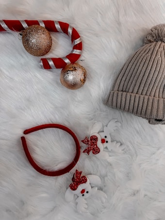 A cozy winter scene featuring a knitted beanie with a pom-pom next to a red and white candy cane decoration with two glittery gold ornaments. A headband with plush white reindeer figures adorned with red bows completes the arrangement, all set on a soft, fluffy white surface.