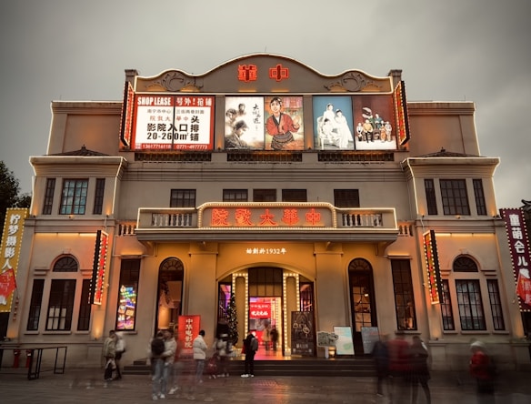 A building with a prominent facade featuring bright lights and electronic billboards. The structure includes multiple lit signs in Chinese characters, illuminated posters, and decorative architectural elements. Several people are walking in front of the bustling entrance, amidst signs advertising a shop lease and various other displays.
