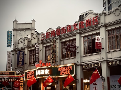 A historic street area with traditional architecture, featuring multiple signs with Chinese characters. The buildings have intricate designs, and there are several red national flags displayed prominently. Neon lights and vintage-style signage suggest a vibrant cultural or commercial hub.