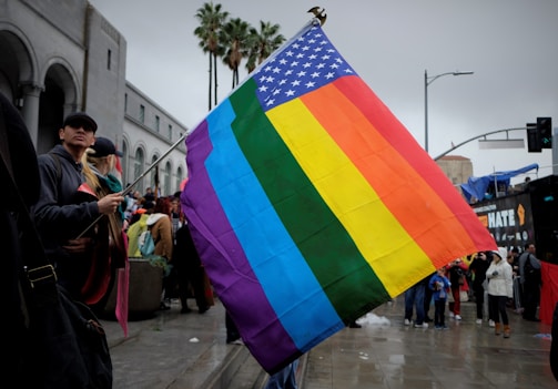 A large rainbow pride flag combined with the American flag is prominently displayed in the foreground. A crowd of people stands nearby on an overcast day, likely part of a public gathering or protest. In the background, the architecture of a building with arches is visible along with some palm trees.