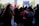 A crowd of people gather at a rally, with several holding signs. A young person with red hair stands in the foreground, looking to the side. The atmosphere is busy with individuals engaging in conversations, some capturing photographs. The signs include messages of empowerment and social justice.