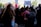 A crowd of people gather at a rally, with several holding signs. A young person with red hair stands in the foreground, looking to the side. The atmosphere is busy with individuals engaging in conversations, some capturing photographs. The signs include messages of empowerment and social justice.