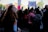 A crowd of people gather at a rally, with several holding signs. A young person with red hair stands in the foreground, looking to the side. The atmosphere is busy with individuals engaging in conversations, some capturing photographs. The signs include messages of empowerment and social justice.