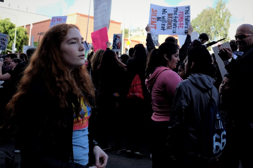 A crowd of people gather at a rally, with several holding signs. A young person with red hair stands in the foreground, looking to the side. The atmosphere is busy with individuals engaging in conversations, some capturing photographs. The signs include messages of empowerment and social justice.