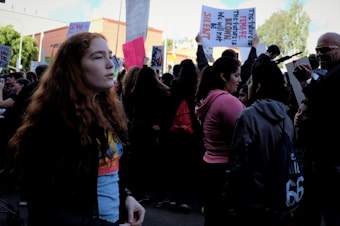 A crowd of people gather at a rally, with several holding signs. A young person with red hair stands in the foreground, looking to the side. The atmosphere is busy with individuals engaging in conversations, some capturing photographs. The signs include messages of empowerment and social justice.