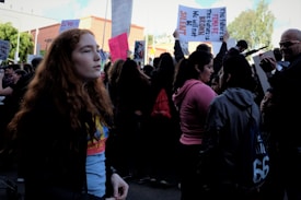 A crowd of people gather at a rally, with several holding signs. A young person with red hair stands in the foreground, looking to the side. The atmosphere is busy with individuals engaging in conversations, some capturing photographs. The signs include messages of empowerment and social justice.