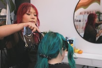 A person with red hair, wearing a black outfit and silver jewelry, is styling another person's teal hair inside a salon. The scene is reflected in a round mirror on the wall, and there are small decorative pumpkins on a white counter in the background.