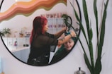 A hairstylist with red hair is dyeing another person's hair green, as seen in the reflection of a circular mirror. The room has a colorful wave pattern on the wall and various products and plants on shelves in the background.