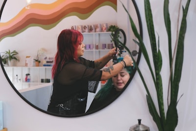 A hairstylist with red hair is dyeing another person's hair green, as seen in the reflection of a circular mirror. The room has a colorful wave pattern on the wall and various products and plants on shelves in the background.