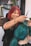 A hairstylist with red hair and tattoos is smiling while working on a client's vibrant teal hair. The setting is a modern salon with plants and shelves in the background.