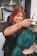 A hairstylist with red hair and tattoos is smiling while working on a client's vibrant teal hair. The setting is a modern salon with plants and shelves in the background.